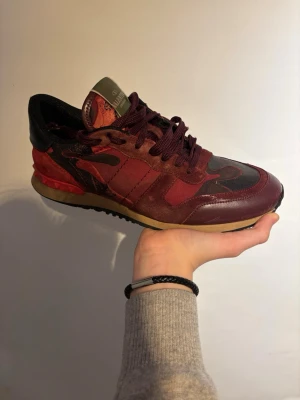 Valentino Rockrunner sneakers vinröd - Snygga Valentino Rockrunner sneakers i vinrött och svart med coola camouflagemönster. Skorna har snörning, rund tå och detaljer i mocka, skinn och textil. Sulan är beige med svarta inslag och baksidan har de ikoniska nitarna.