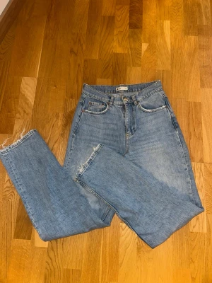 Ljusblåa raka jeans - Säljer ett par klassiska blå jeans från Gina Tricot i modell med hög midja och raka ben. Jeansen har fickor, dragkedja och knapp framtill samt råa, fransiga benslut för en avslappnad vibe. Perfekta till sneakers eller boots.