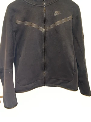 Svart Nike Tech XS/S - Säljer en svart Nike hoodie i storlek XL (barn) och passar ungefär som en size XS-S i vanliga storleker. Den har hel dragkedja och hög krage. Tröjan har diskreta svarta detaljer över bröstet och Nike-logga på vänster sida. Tillverkad i mjukt material, perfekt för chill dagar eller träning. 