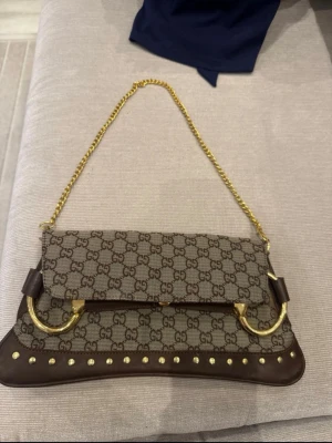 Brun monogram axelväska med kedja - Snygg axelväska i brunt med klassiskt GG-mönster. Väskan har guldfärgade detaljer, kedjerem och nitar längs kanterna. Materialmix av skinn och canvas, med stora guldfärgade ringar som dekoration. Perfekt för dig som vill sticka ut med en ikonisk design., har slitningar på insidan men inget som försvårar användningen🫶🏻