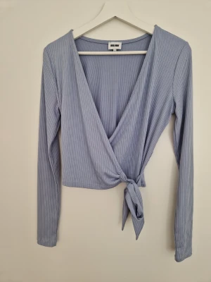 Ljusblå omlottopp från Bubbleroom - Säljer en ljusblå omlottopp från Bubbleroom i ribbad trikå. Toppen har långa ärmar, djup v-ringning och knyt i sidan för en snygg passform. Perfekt till jeans eller kjol för en fräsch look.