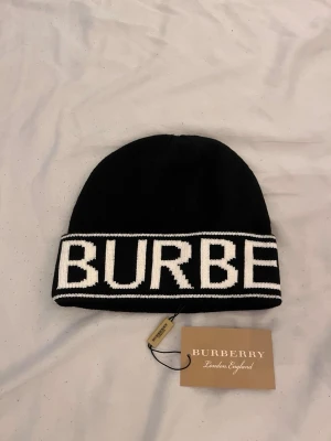 Svart mössa från Burberry - Snygg svart mössa från Burberry med bred vikt kant där BURBERRY står i stora vita bokstäver. Klassisk och stilren design, perfekt för dig som vill ha en exklusiv accessoar. Stickad i mjukt material för skön känsla och passform.