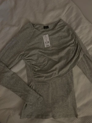 Grå långärmad topp från Gina Tricot - Snygg grå långärmad topp från Gina Tricot med lätt rynkad detalj på sidan. Tillverkad i mjuk bomullsmix och har en figurnära passform. Perfekt att styla med jeans eller kjol för en clean look.