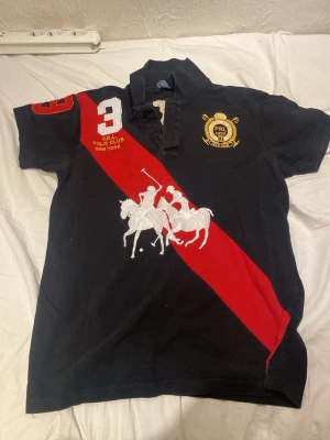 Svart pikétröja från Polo Ralph Lauren - Svart pikétröja från Polo Ralph Lauren med röd diagonal rand, stor broderad polospelare på framsidan och guldfärgat emblem på bröstet. Klassisk krage, knappslå och siffran 3 på axeln. Perfekt för en sportig och stilren look.