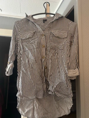Randig skjorta med knappar - Snygg skjorta med svartvita ränder, två stora bröstfickor och knappar framtill. Skjortan har lång ärm som kan vikas upp och fästas med knapp. Lätt och luftig modell med längre bakstycke, perfekt till jeans eller kjol. Använd en gång💞