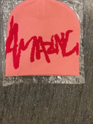 Rosa mössa med röd grafisk text - Säljer en rosa mössa med röd grafisk text framtill i streetstil. Mössan är slätstickad och har en mjuk, stretchig passform. Perfekt accessoar för att addera färg till din outfit.