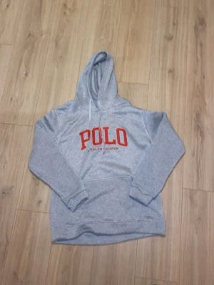 Grå hoodie från Polo Ralph Lauren - Snygg grå hoodie från Polo Ralph Lauren med stor röd POLO-logga på bröstet och klassisk känguruficka. Tröjan har huva med vita snören och ribbade muddar vid ärmar och nederkant. Perfekt för en avslappnad och trendig look. Den är från CNfans därav lågt pris. 