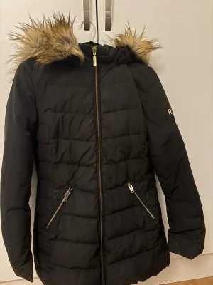 Svart dunjacka med fuskpäls från Rockandblue - Svart quiltad dunjacka från Rockandblue med huva och fuskpälsdetalj. Jackan har dragkedja framtill, två snedställda fickor med dragkedja och en snygg logga på ärmen. Perfekt för kalla dagar och riktigt skön passform. Märks lite att den är använd men inte alls mycket 