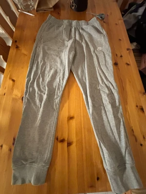 Grå mjukisbyxor från Nike - Säljer ett par grå mjukisbyxor från Nike med elastisk midja och muddar vid bensluten. Byxorna har en enkel och clean design med Nike-logga på låret. Perfekta för chill eller träning.