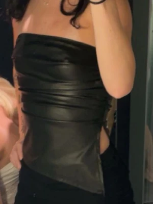 Svart skinn topp - Säljer en svart topp i skinnimitation. Toppen är axelbandslös och har en tight passform med en cool, glansig finish. Perfekt för dig som vill sticka ut med en edgy look.