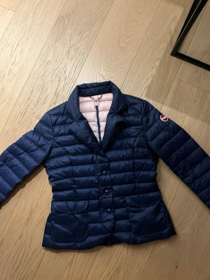 Colmar Originals Lättdunjacka / Blazer – Marinblå – Stl 38 (IT 44) - ❤️❤️❤️Snygg blå pufferjacka från Colmar med quiltad design och klassisk logga på ärmen. Jackan har tryckknappar framtill, två fickor och en hög krage. Materialet är glansigt och lätt, perfekt för kyliga dagar. Insidan är ljusrosa med Colmar-etikett.❤️❤️❤️❤️❤️❤️❤️
