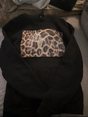 Svart hoodie med elefant och leopardmönster - Cool svart hoodie med en stor patch på bröstet som har leopardmönster och en vit elefant ovanpå. Klassisk huva med snörning och magficka framtill. Perfekt för dig som gillar djurmotiv och streetwear vibes.
