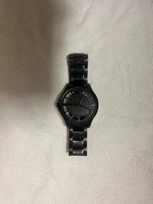 Svart klocka från Armani Exchange - Stilren svart klocka från Armani Exchange med metallarmband och rund urtavla. Urtavlan har ett rutigt mönster i mitten och tydliga indexmarkeringar. Klockan har en modern och minimalistisk design som passar till många olika stilar.