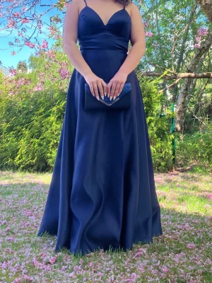 Marinblå långklänning med snörning - Elegant marinblå långklänning med smala axelband, v-ringning och snygg snörning i ryggen. Klänningen har en figurnära överdel och ett utsvängt, svepande kjolparti. Perfekt för bal eller festliga tillfällen. Storlek 40/42