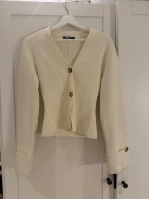 Krämvit kofta från Gina Tricot - Stilren krämvit kofta från Gina Tricot med v-ringning och två stora bruna knappar framtill. Koftan har långa ärmar med dekorativa knappar vid ärmsluten och en något croppad passform. Perfekt att slänga över en topp för en enkel men snygg look.