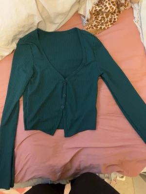 Mörkgrön ribbad tröja - Snygg mörkgrön ribbad tröja med v-ringning och tre knappar framtill. Modellen är croppad och har långa ärmar.
