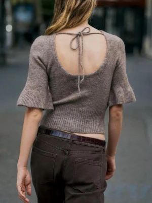 Beige stickad topp med öppen rygg - Ny med prislapp. Supersöt beige stickad topp med korta trumpetärmar och öppen rygg som knyts med en rosett i nacken. Toppen har ribbade detaljer i midjan och vid ärmsluten, vilket ger en snygg passform och trendig look.