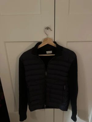 Svart cardigan från Moncler - Snygg svart cardigan från Moncler med quiltad front och dragkedja. Jackan har långärmade ärmar i mjukt material och klassisk Moncler-logga på ärmen. Perfekt för dig som vill ha en stilren och trendig look.