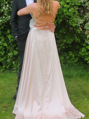 Lång champagnefärgad balklänning - Elegant långklänning i champagnefärgad satin med smala axelband och öppen rygg med snörning. Klänningen har en v-ringad front och ett svepande fall som ger en lyxig känsla. Perfekt för bal eller speciella tillfällen.