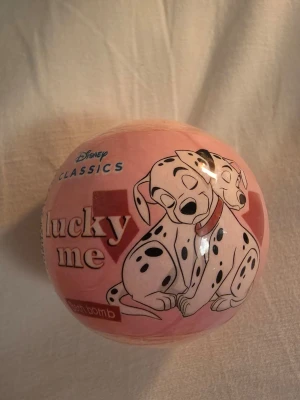Disney Lucky Me badbomb 101 dalmatiner - Söt badbomb från Disney Classics med motiv av 101 dalmatiner och texten 'lucky me'. Badbomben är rosa och väger 100g. Perfekt för ett mysigt bad och ger en rolig touch till badstunden.