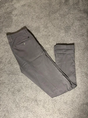 Grå chinos från Mason's - Snygga grå chinos från Mason's med klassisk passform och diskreta detaljer. Byxorna har en bakficka med knapp och är tillverkade i ett mjukt bomullsmaterial som känns bekvämt hela dagen. Perfekta för en clean och stilren look.