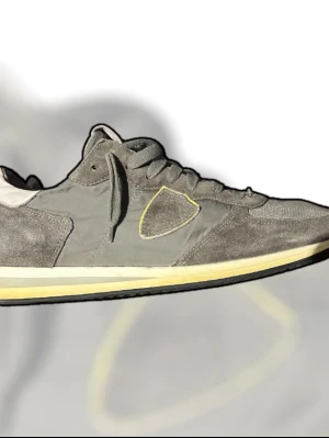 Grå Philippe Model sneakers med gul sula - Snygga sneakers från Philippe Model i olika nyanser av grått med detaljer i mocka och mesh. Skorna har snörning, vit och gul sula samt en ikonisk sköld på sidan med gul kant. Perfekt för dig som gillar stilrena och sportiga skor.