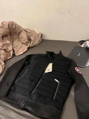 Svart dunväst från Canada Goose - Snygg svart dunväst från Canada Goose med quiltad framsida, två dragkedjeförsedda fickor och klassisk logopatch på ärmen. Västen har hög krage och stilren design som passar perfekt till lager-på-lager. Dragkedja framtill och diskreta detaljer.