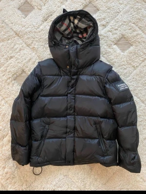Svart pufferjacka från Burberry - Snygg svart pufferjacka från Burberry med klassiskt rutigt foder i huvan och patch med logga på ärmen.  Helt ny bara utan prislapp använd bara testad priset kan sänkas