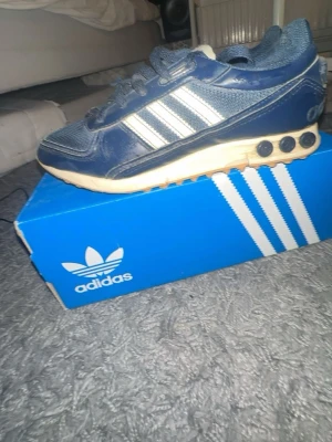 Blå Adidas sneakers med vita ränder - Snygga blå sneakers från Adidas med klassiska vita ränder på sidan och meshdetaljer framtill. Skorna har snörning och en beige sula med svarta detaljer. Perfekta för dig som gillar retrostil och sportig look.