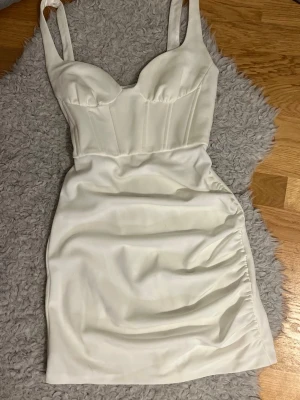 Vit kort klänning - Snygg vit kort klänning med markerad byst. Klänningen har draperad detalj på sidan och figurnära passform. Perfekt för dig som vill ha en stilren och trendig look. Materialet känns mjukt och stretchigt. Klänningen är i storlek M men passar mig som brukar ha på mig storlek S och Xs. 