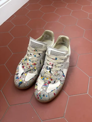 Maison Margiela sneakers med färgstänk - Unika sneakers från Maison Margiela i vitt och beige med färgstänk i flera färger. Skorna har snörning, rund tå och platt sula. Tillverkade i skinn och mocka för en lyxig känsla och cool look. Använd skick och jag har rivit av färg på dem vita delarna längst fram på skorna 