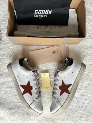 Golden Goose vita sneakers med röd stjärna - Golden Goose sneakers i vitt skinn med ikonisk röd stjärna på sidan och grå snörning. Skon har en beige platt sula och svart detalj på hälen. Klassisk och stilren modell som passar till det mesta.