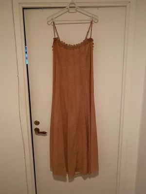 Beige långklänning med smala axelband - Luftig beige långklänning med smala axelband och volangdetalj upptill. Klänningen har en avslappnad passform och ett diskret rutigt mönster. Knytband i ryggen för justerbar passform. Perfekt för varma dagar.