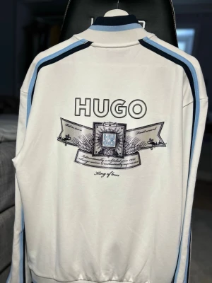Vit ziptröja med blå detaljer HUGO -  Tröjan går inte att köpa längre! Helt äkta! Vit ziptröja från HUGO med blå och mörkblå ränder längs ärmarna. Stor logga och grafiskt tryck på ryggen samt mindre emblem på bröstet. Tröjan har hög krage, dragkedja framtill och två fickor med dragkedja. Perfekt för en sportig och trendig look. 