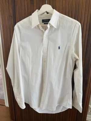 Vit skjorta från Polo Ralph Lauren - Klassisk vit herrskjorta från Polo Ralph Lauren i slim fit-modell. Skjortan har lång ärm, knappar framtill och den ikoniska blå broderade loggan på bröstet. Tillverkad i mjuk bomull för en clean och stilren look.