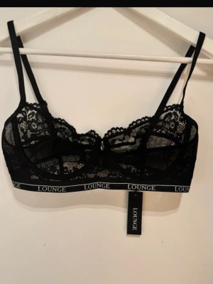 Svart spets-bh från Lounge - Säljer en svart bh från Lounge med tunna axelband och bred resår med logga. Bh:n är i genomskinlig spets med blommigt mönster och har bygel för extra stöd. Perfekt för dig som gillar en stilren och trendig look.