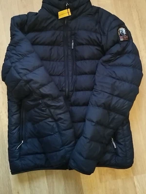 Svart dunjacka från Parajumpers - Säljer en svart dunjacka från Parajumpers med klassisk patch på ärmen och gul detalj vid kragen. Jackan har quiltad design, dragkedjor på fickorna och hög krage. Insidan är ljusgrå och materialet är lätt och smidigt, perfekt för kyliga dagar.