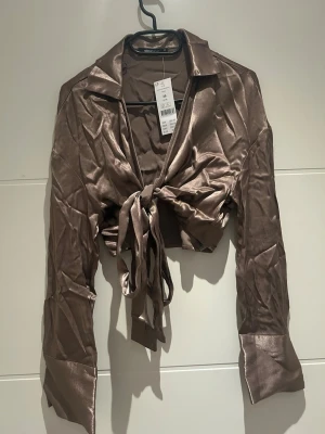 Brun omlottopp med knyt från Gina Tricot - Snygg brun omlottopp från Gina Tricot i glansigt material. Toppen har långa ärmar, krage och knyt framtill som ger en croppad look. Perfekt för dig som vill ha en trendig och stilren topp med lite extra attityd. Helt ny.