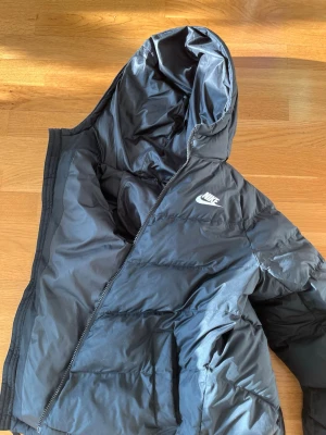 Svart pufferjacka från Nike - Säljer en svart pufferjacka från Nike med huva och dragkedja. Jackan har en quiltad design och Nike-logga på bröstet. Perfekt för kalla dagar och riktigt skön att bära. Passar dig som gillar en sportig och clean stil.