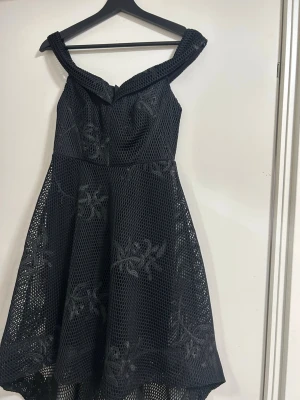 Svart offshoulder klänning med spets - Snygg svart offshoulder klänning med mesh och broderade blommor. Klänningen har en markerad midja, vidd i kjolen och dragkedja bak. Materialet är mesh med spetsdetaljer som ger en cool och elegant look.