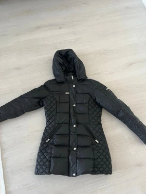 Svart dunjacka Rockandblue quiltad - Snygg svart dunjacka från Rockandblue med quiltade partier på sidorna och ärmarna. Jackan har huva, dragkedja och knappar framtill samt flera fickor med dragkedja. Märkespatch på ena ärmen och figurnära passform. Perfekt för kalla dagar! 36 men passar även mig som bär 34 i vanliga fall.