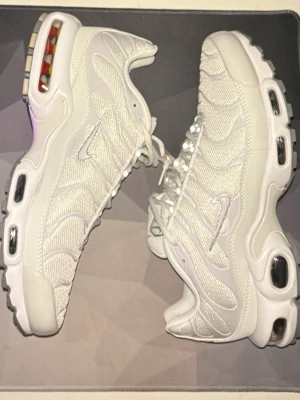 Nike TN vit - Nike Air Max Plus sneakers i helvitt med mesh och syntetpaneler. Skorna har tydliga Air Max-bubblor i sulan, reflekterande detaljer och klassisk snörning. Swoosh-logga på sidan och AIR-märke på plösen. Perfekta för dig som gillar stilrena och sportiga sneakers.