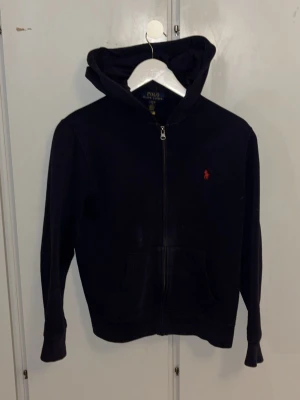 Svart hoodie från Polo Ralph Lauren - Snygg svart hoodie från Polo Ralph Lauren med dragkedja framtill och klassisk röd logga broderad på bröstet. Hoodien har huva och kängurufickor, tillverkad i mjukt bomullsmaterial. Perfekt för en avslappnad och stilren look. Den är storlek L (14-16 år) men sitter som XS/S