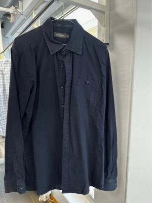 Mörkblå skjorta från Dressmann L - Klassisk mörkblå skjorta från Dressmann i storlek L. Skjortan har lång ärm, bröstficka och knappar framtill. Tillverkad i 100% bomull för en skön och luftig känsla. Perfekt till jeans eller chinos för en clean look. Solmärken vid kragen.