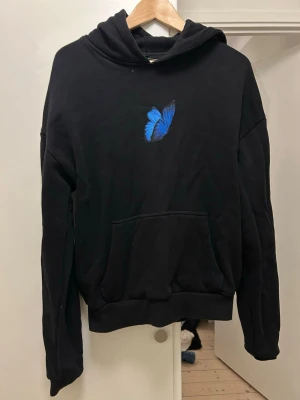 Svart hoodie med blå fjäril Mister Tee - Svart hoodie från Mister Tee med en snygg blå fjäril broderad på bröstet. Klassisk känguruficka framtill och huva. Perfekt för dig som gillar stilrena plagg med en unik detalj. Mjuk och skön bomullsblandning.