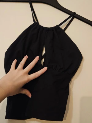 Svart croppad linnetopp med cut-out - Svart croppad linnetopp med smala axelband som korsas i ryggen och en snygg cut-out detalj framtill. Toppen har en halterneck-liknande form.