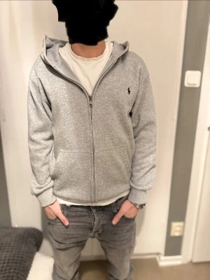 Grå hoodie från Polo Ralph Lauren - Snygg grå hoodie från Polo Ralph Lauren med klassisk logga broderad på bröstet. Är oanvänd, tröjan har luva, dragkedja framtill och två fickor. 