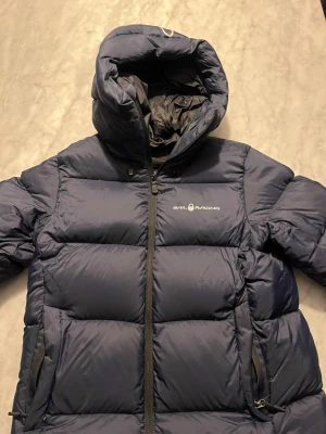 Mörkblå Sail Racing dunjacka L - Säljer en mörkblå pufferjacka från Sail Racing i storlek Large. Jackan har fyllning av högkvalitativt dun (700 fill power), huva, två dragkedjeförsedda fickor och Sail Racing-logga på bröstet och huvan. Perfekt för kalla dagar och riktigt snygg streetstil. Den är i super bra skick och har endast blivit använd 5 gånger max. Pris kan diskuteras!