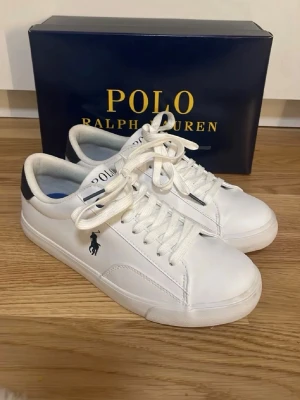Vita sneakers från Polo Ralph Lauren - Stilrena vita sneakers från Polo Ralph Lauren med klassisk logga på sidan och svart detalj vid hälen. Skorna har snörning och är tillverkade i slätt skinnliknande material. Perfekta för en clean och sportig look. Använda endast en gång och säljer då de inte passar 