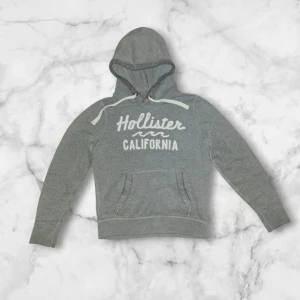 Grå hoodie från Hollister med tryck - Grå hoodie från Hollister med vit text 'Hollister California' på bröstet. Tröjan har huva med snörning och en stor magficka framtill. Mjuk och skön, perfekt för chill dagar. Klassisk passform och långärmad design.